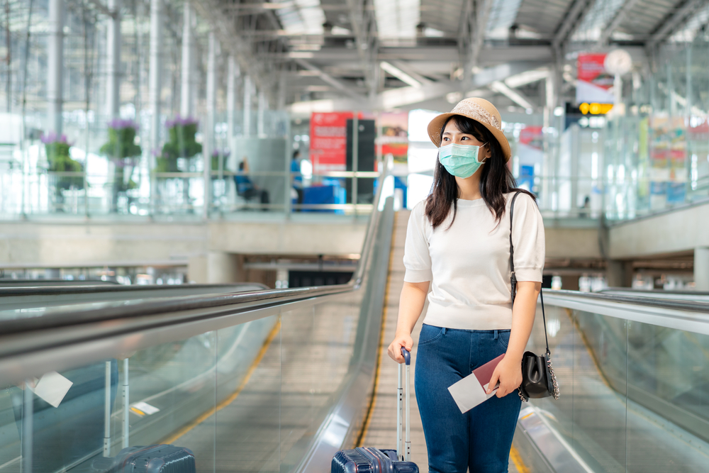 a woman wearing a mask walking in an airport with a rolling suitcase