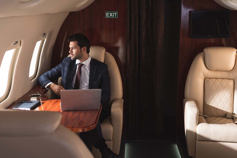 A businessman sits in a seat on a private jet with his laptop open on the table in front of him.