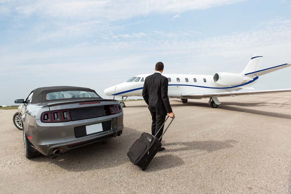 A man with a rolling suitcase steps out of a car and toward a small white plane on the pavement.